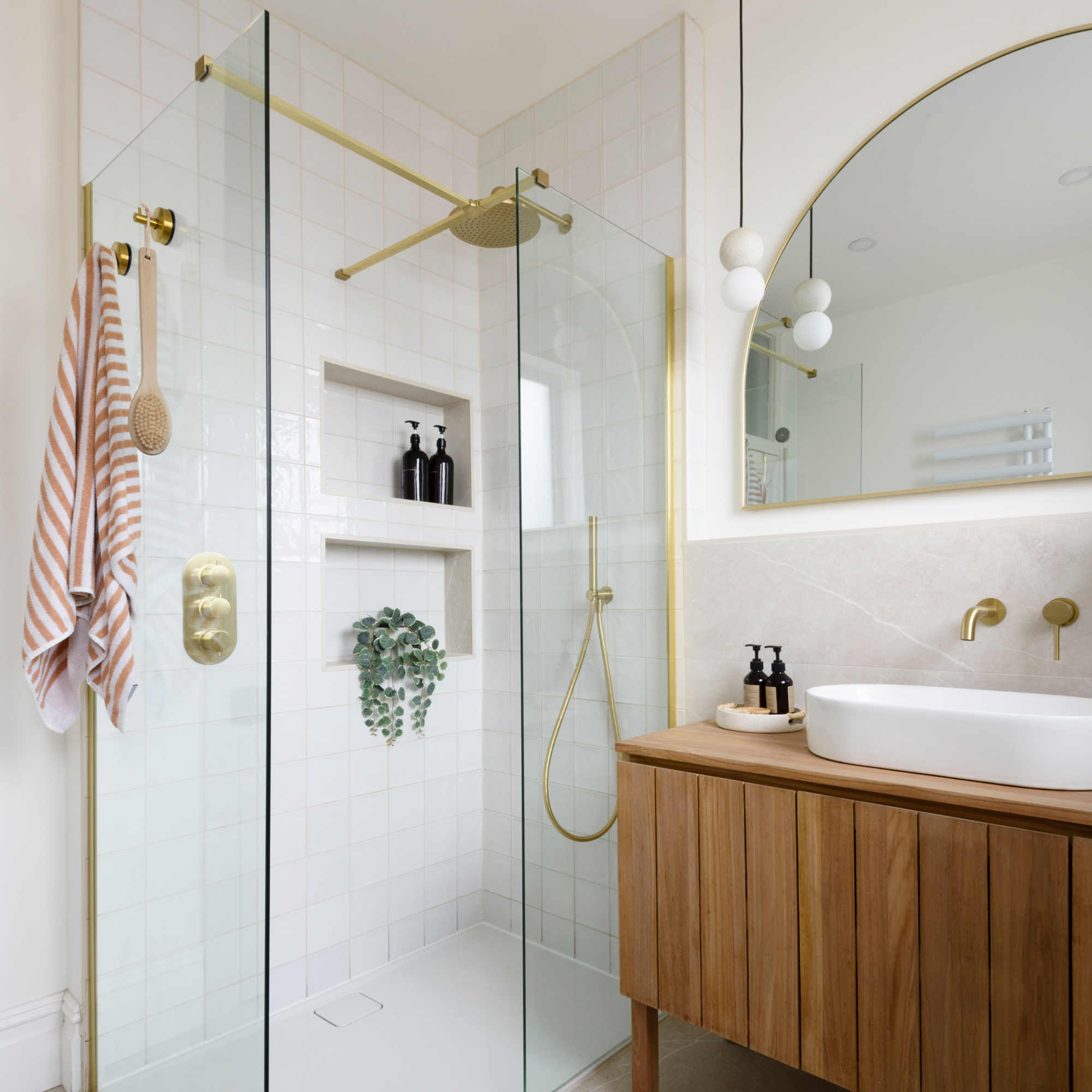 a shower enclosure with glass panels and with pale coloured tiles and brass fixtures, beside a wooden vanity unit and half-moon wall mirror