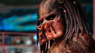 Close up image of Predator, a terrifying alien with a large mouth full of fanged teeth, small yellow eye and dreadlocks.