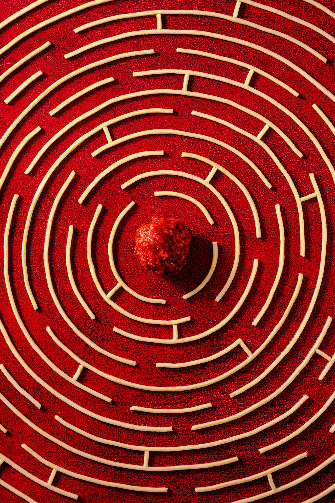 A top-down view of a red spiral maze made of pasta, with a textured red ball in the center, creating a playful and artistic composition
