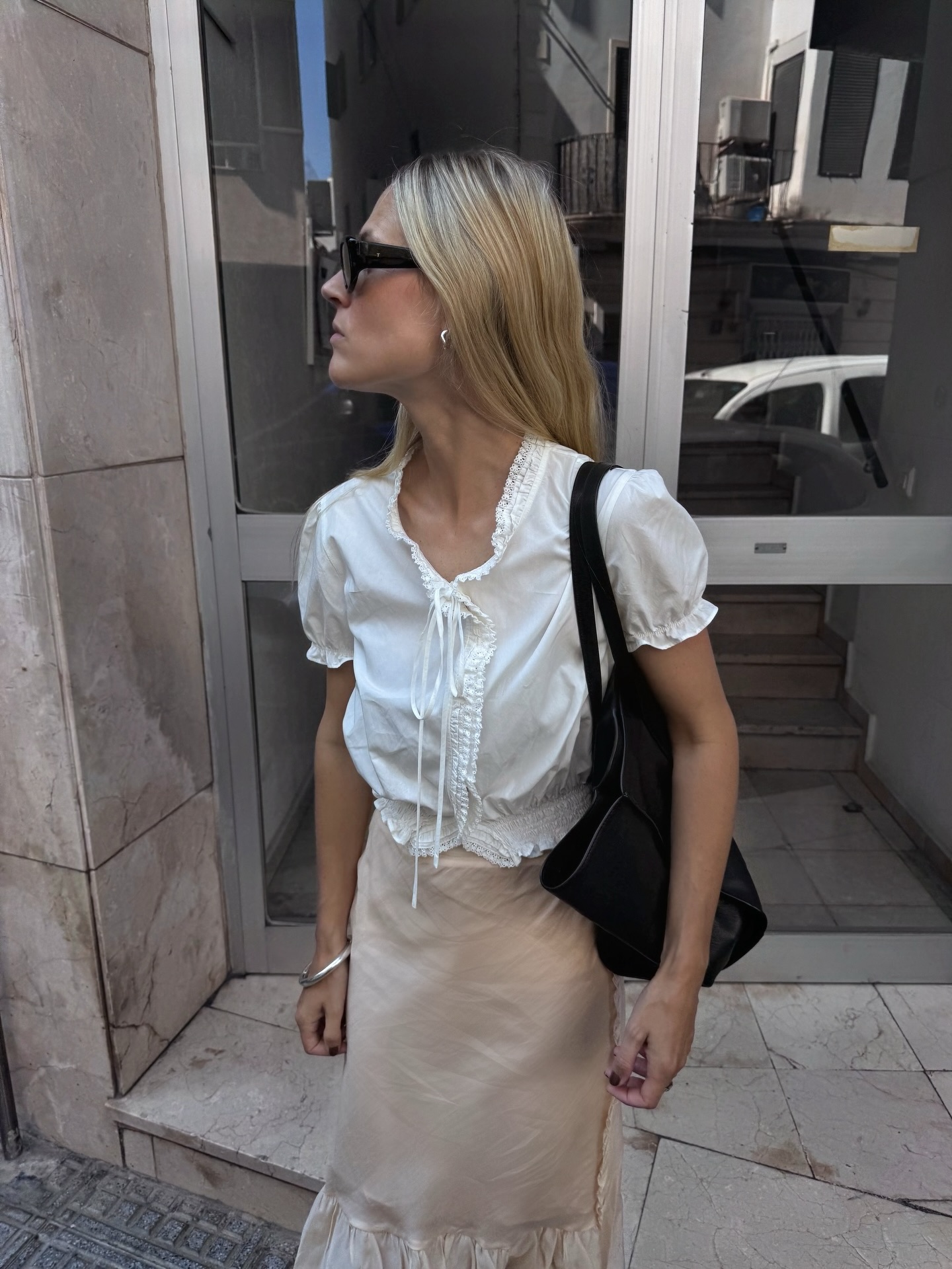 An image of an influencer wearing a white peasant blouse with a beige satin slip skirt and sunglasses standing in front of a building