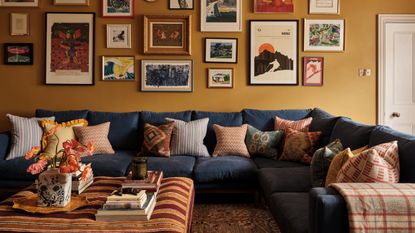 Living room with mustard walls decorated with a collection of prints and paintings, a large dark blue sectional sofa with patterned pillows scattered around it, and a striped ottoman at the centre.