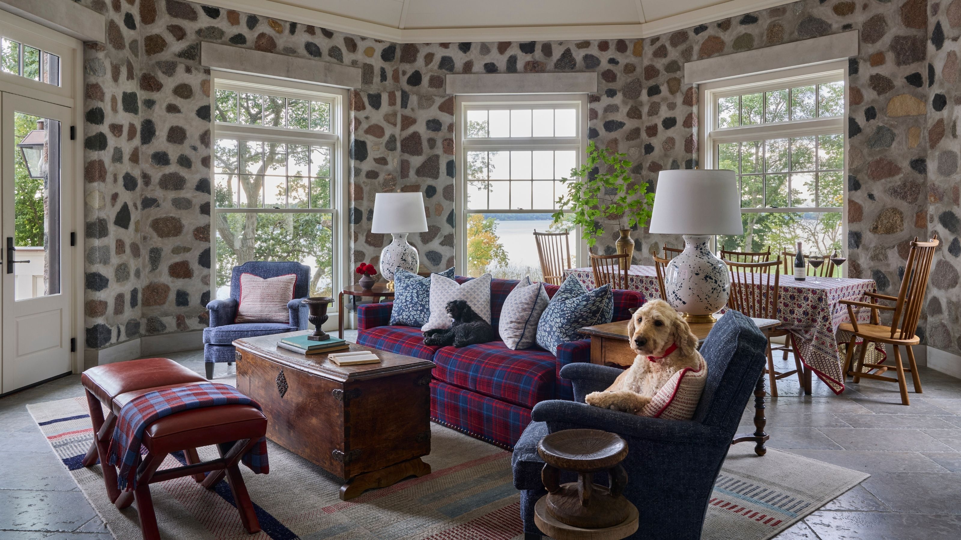 Sunroom featuring exposed fieldstone walls, large grid windows, a red-and-blue plaid sofa, blue accent armchair, and a golden retriever dog resting comfortably in a armchair.