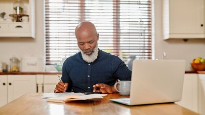 An older man uses a calculator while working on paperwork with a laptop on his kitchen table.