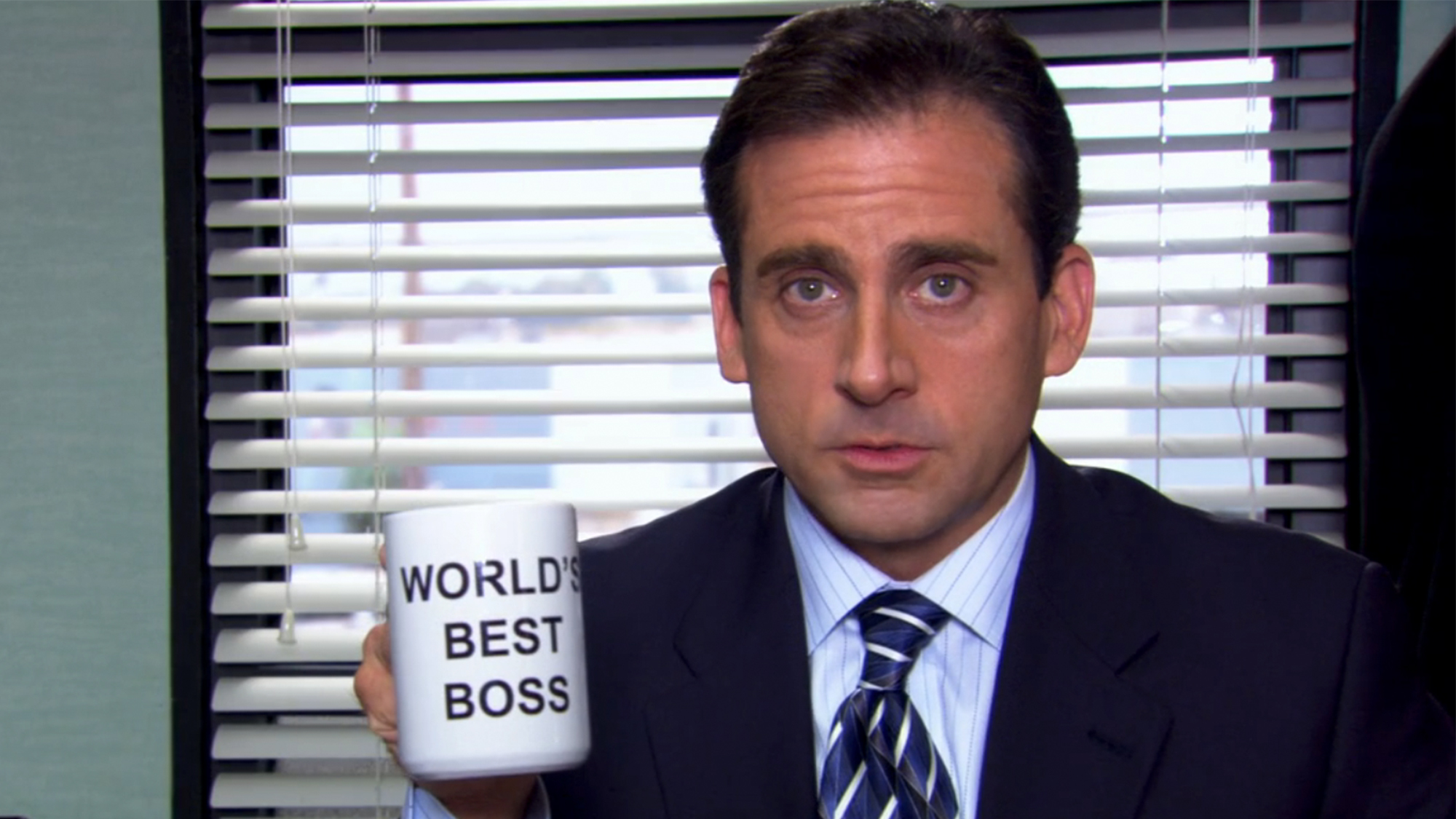 Steve Carell in a suit, holding a coffee mug that says "World's Best Boss" in The Office