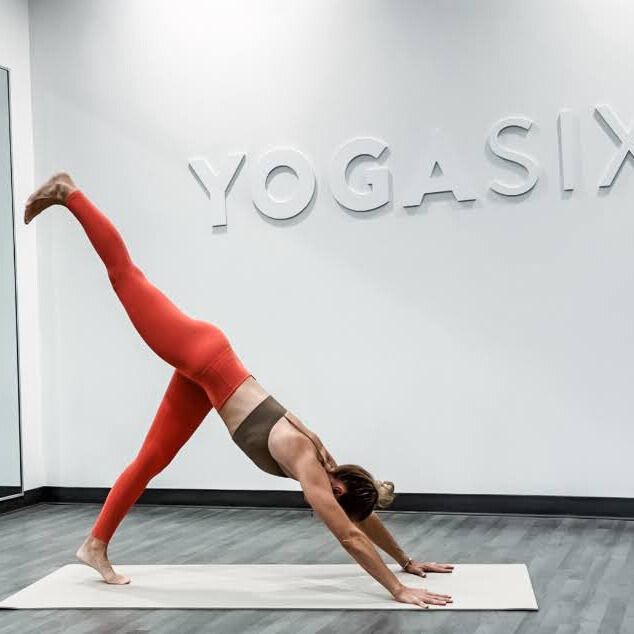 Woman demonstrates yoga pose in yoga studio with words Yoga Six embossed on wall behind her