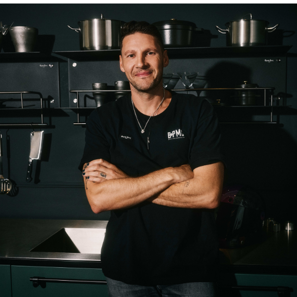 a headshot of Massimo Buster Minale, a man smiling at the camera with his arms crossed wearing a black t-shirt and a necklace, standing in front of pots and pans on kitchen shelving