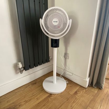 The Silentnight Airmax Pro360 Pedestal Fan in pedestal mode in the corner of a white room, next to a vertical radiator, standing on a wood-look floor