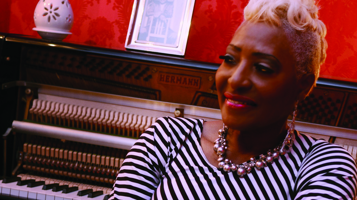 Martha High in a black-and-white stripy top, sitting in front of a piano.