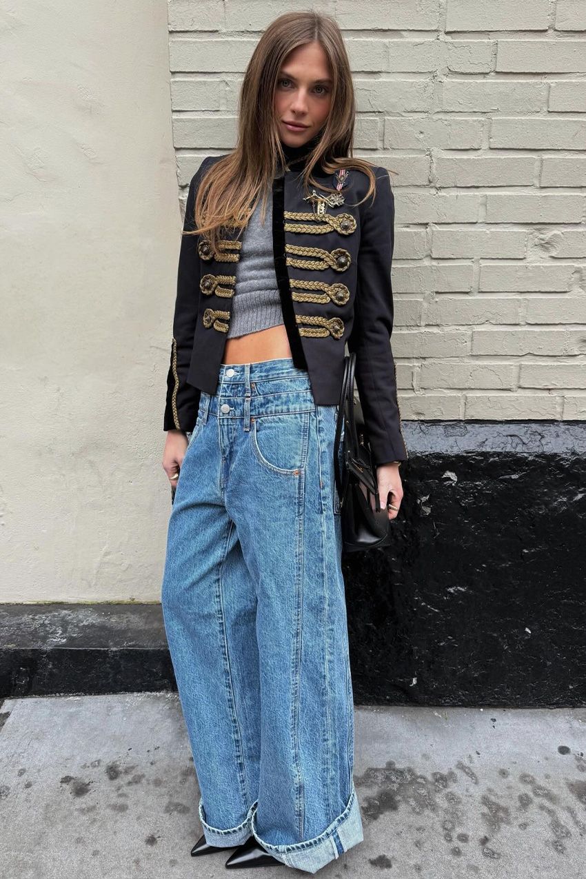 A street style photo of a woman standing outside leaning against a white brick wall wearing a navy blue military jacket with a gray sweater, wide-leg cuffed jeans, and black pointed pumps.