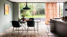 Dining room with table and chairs on patterned rug, picture window glass wall partially covered by patterned orange curtains, leading into and revealing patio garden.