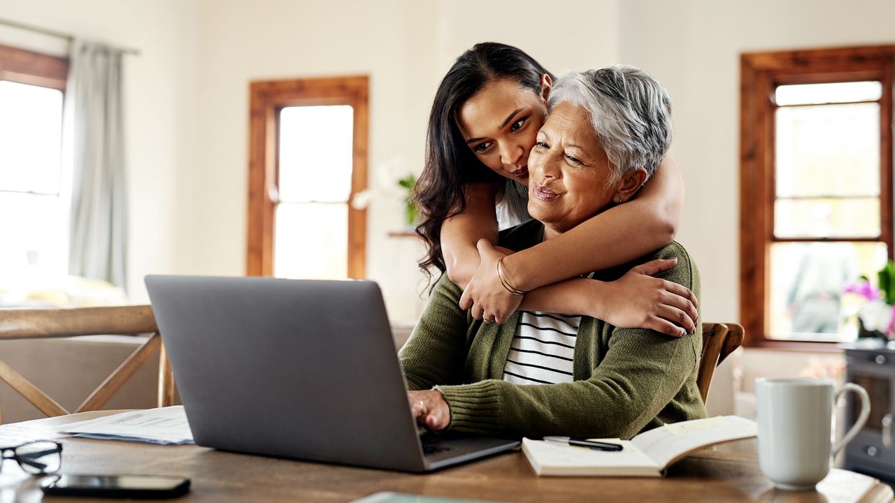 A young woman hugs her mother from behind while they look at a laptop on the dining room table.