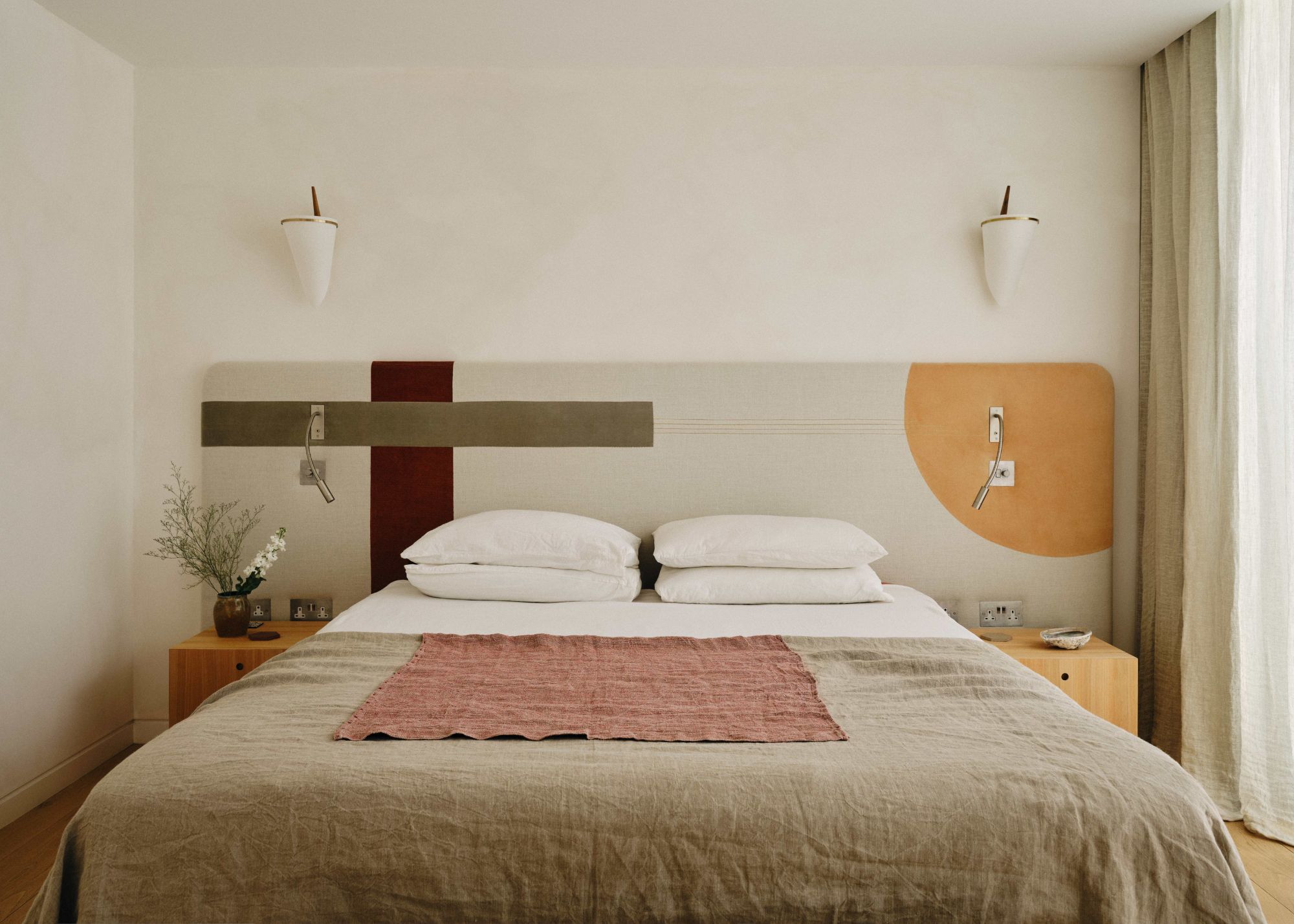A neutral, modern bedroom with warm white walls, a taupe elongated headboard, and bed linens, with mustard, khaki, and dark red patterned shapes on the headboard.