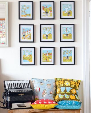 A cream landing with a gallery wall of posters in black frames, and underneath a bench with colourful cushions and a Scarlatti piano accordian