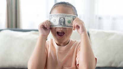 A young girl holds a dollar bill over her eyes.