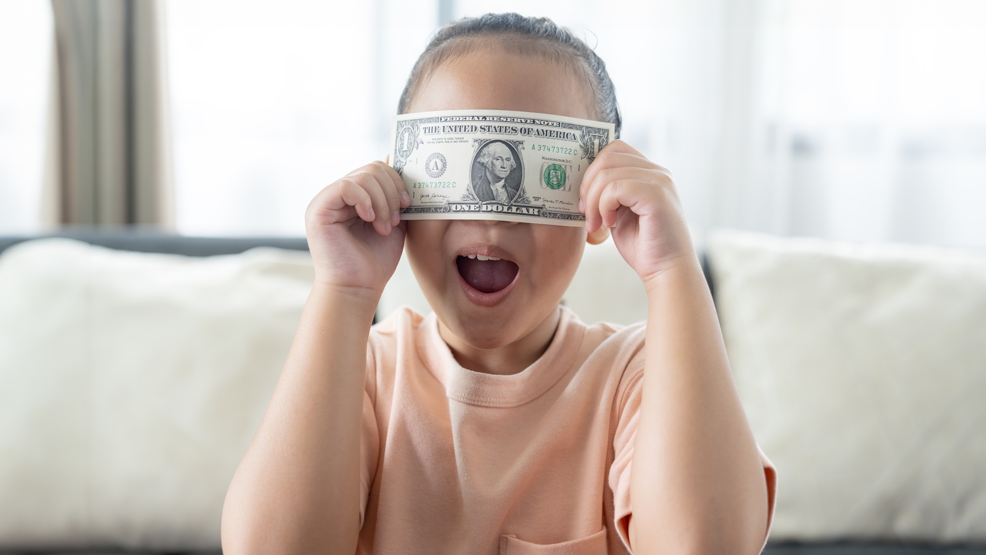 A young girl holds a dollar bill over her eyes.