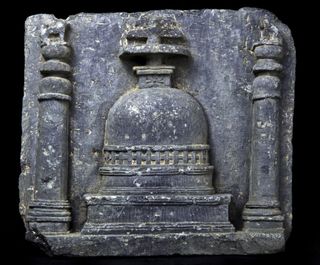 This sculpture shows a stupa, or a place of meditation. A platform called a harmika can be seen near the top of the image. This platform is decorated with a rosette design. Above the harmika, there are three parasol-shaped structures called chattras that