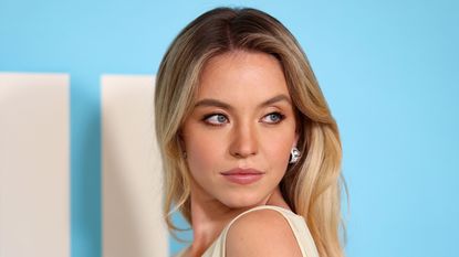 A picture of Sydney Sweeney, a woman with blonde hair wearing a cream colored dress looking over her shoulder, in front of a light blue background