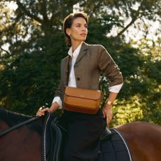 A woman riding a horse, wearing a light brown satchel from Aspinal of London. 