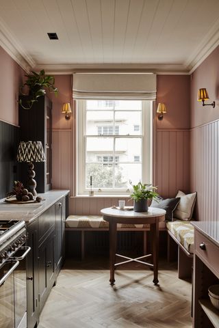 galley kitchen with seated window seat area and small table by window, coral walls and shiplap, black cabinetry, herringbone floor, wall lights, blind