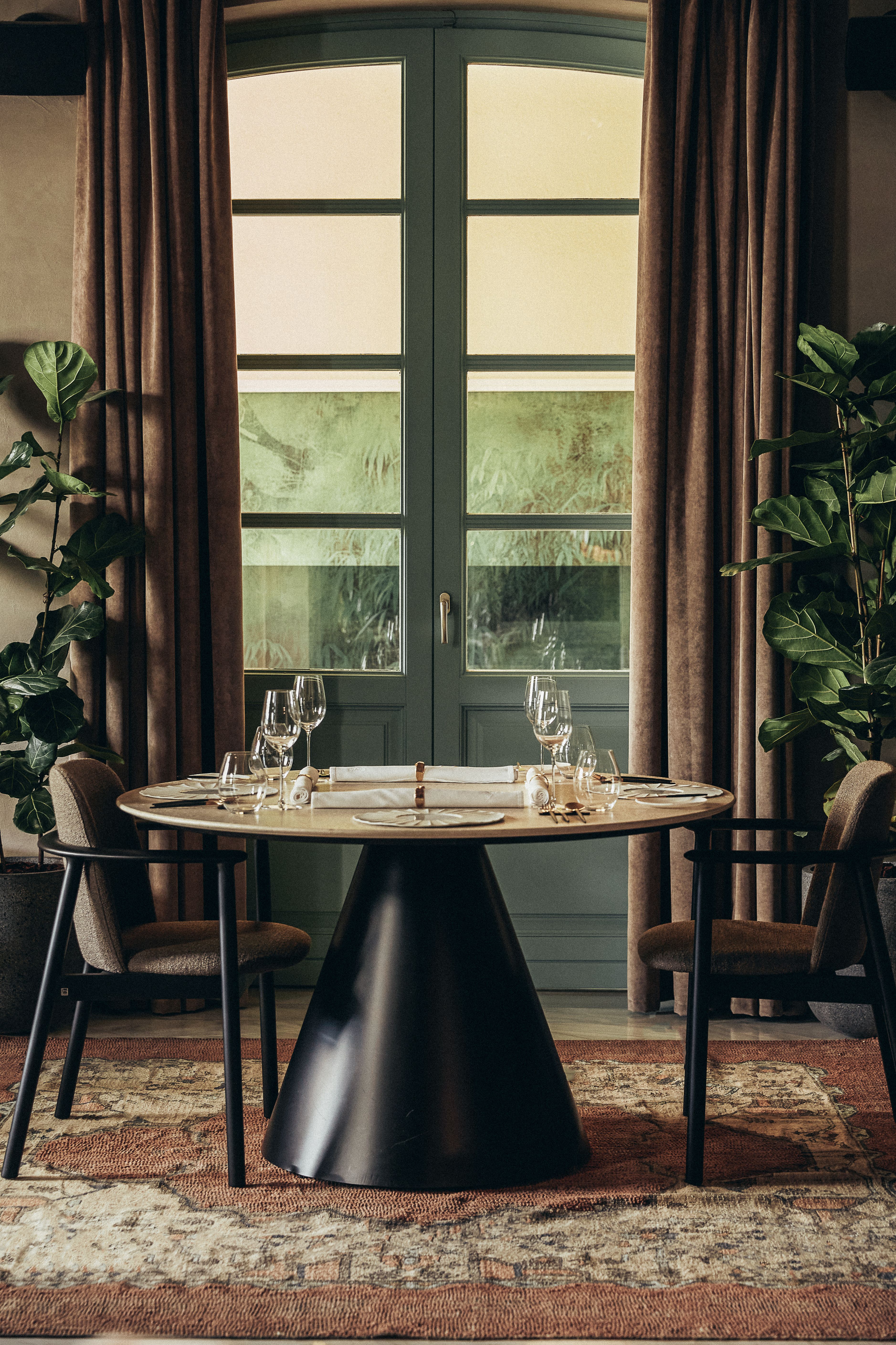 A series of filled red wine glasses against a white background and a contemporary restaurant dining room filled with plant, textured rugs, and slanted furniture in wood.