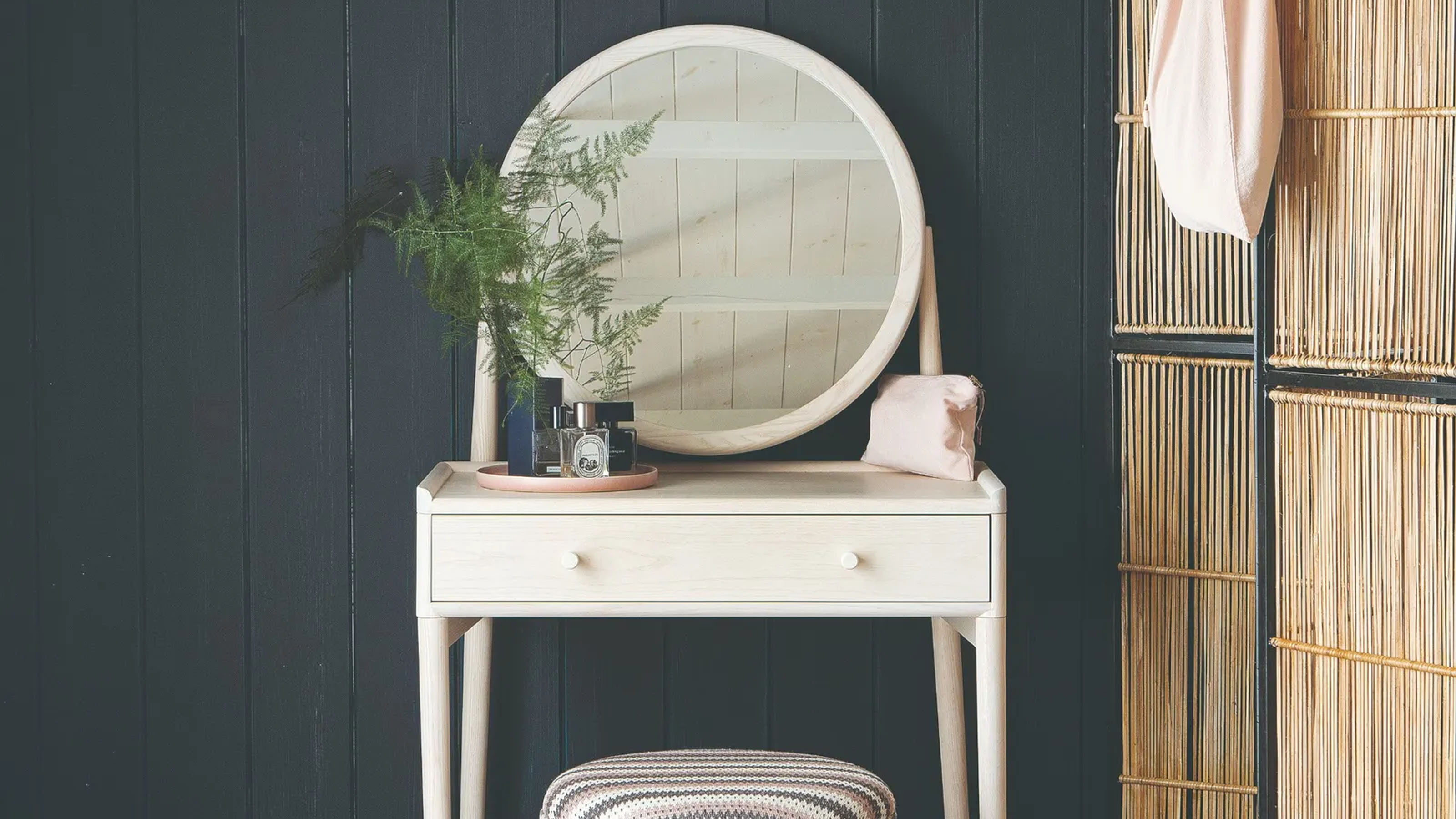 A white dressing table with a round mirror in a room with black walls and a bamboo screen on the right