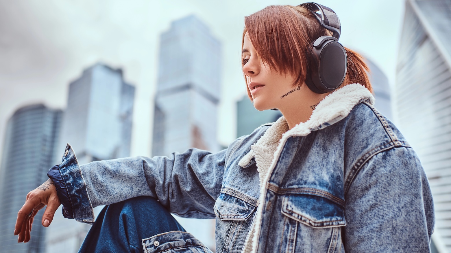 A hipster girl with tattoo on her face trendy dressed wearing headphones listening to music in front of skyscrapers in Moscow city at cloudy morning.