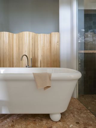 Image of a bathroom with a stand alone white tub that has bun feet. The floor is made of a terracotta-colored marble, and there is a wavy wooden room divider behind the tub.