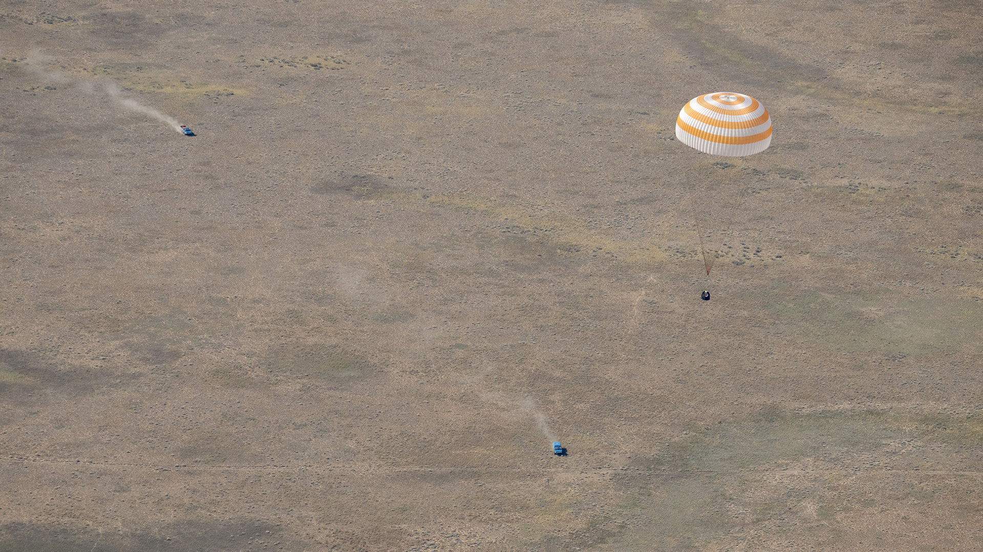 uma fotografia aérea de uma cápsula espacial descendo sob um grande pára-quedas branco e laranja até pousar em uma estepe árida enquanto os veículos de recuperação se aproximam.
