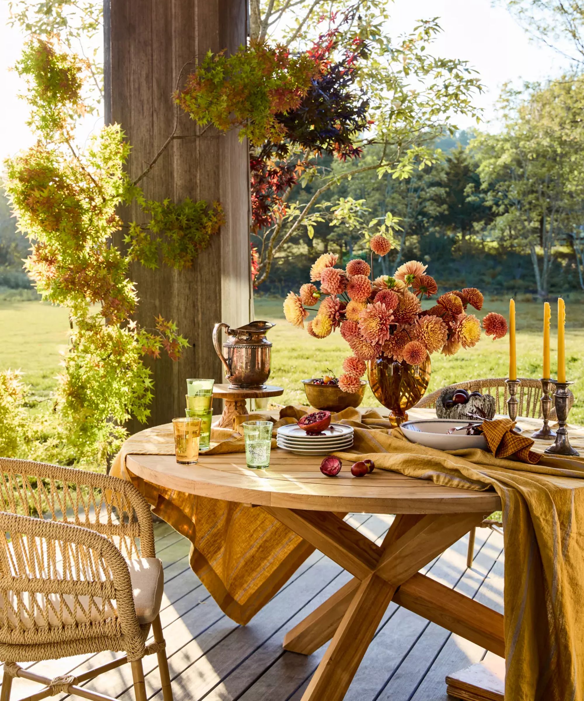 outdoor wooden round table on a porch styled for thanksgiving or fall with dark yellow and orange flowers, linen tablecloth, colored glassware and candlesticks