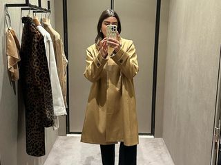 female fashion influencer Anna Newton of The Anna Edit poses for a mirror selfie in a fitting room wearing a trench coat and black pants