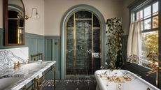 A sophisticated bathroom with muted green wall panelling, off-white walls, a marble double vanity with brass fixtures, an enclosed shower with an arched doorway, and a freestanding bath. 
