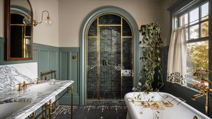 A sophisticated bathroom with muted green wall panelling, off-white walls, a marble double vanity with brass fixtures, an enclosed shower with an arched doorway, and a freestanding bath. 