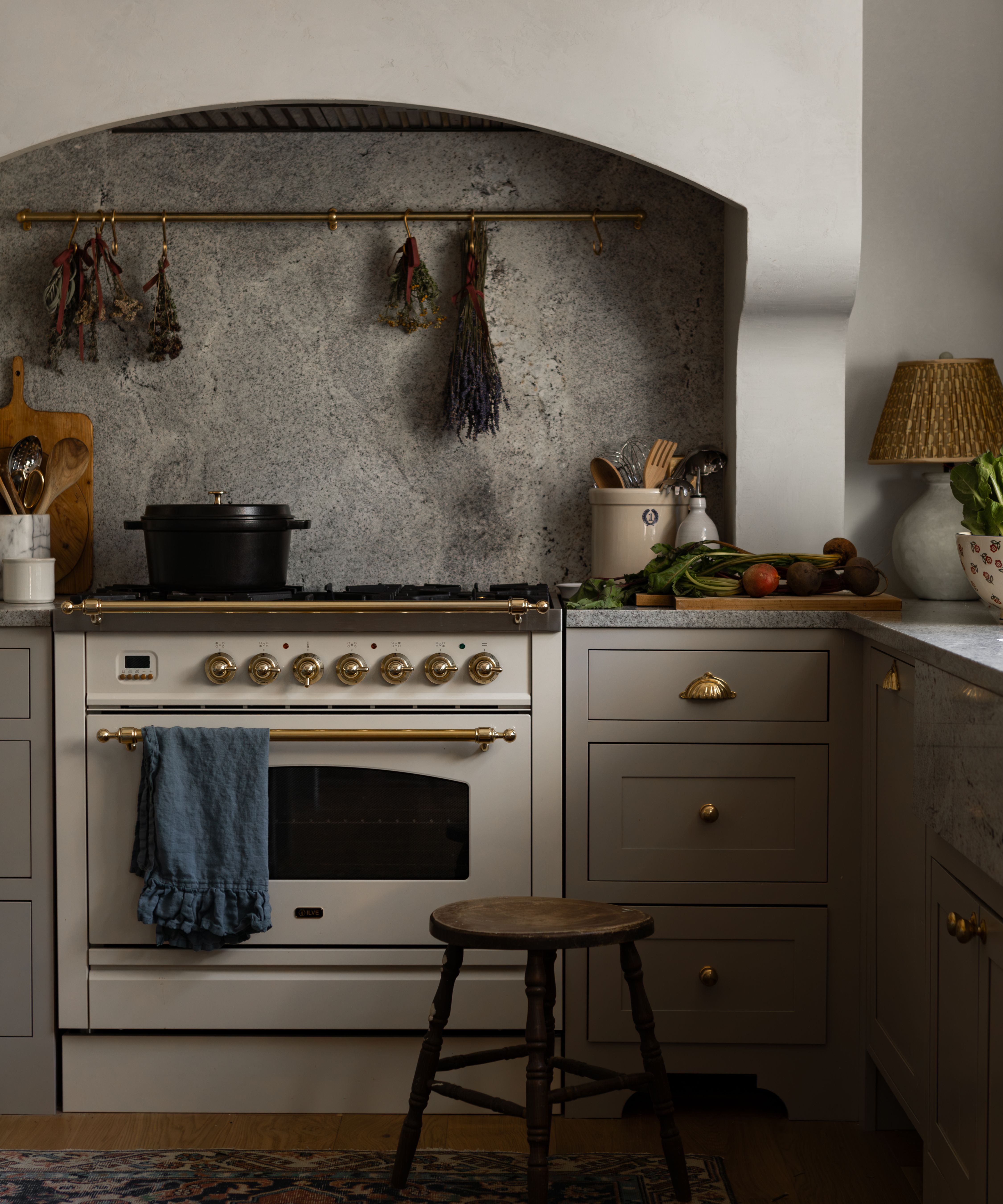 a tiny beige gray kitchen with a plaster hood, small range cooker, marble counters and backsplash with a brass hanging pole