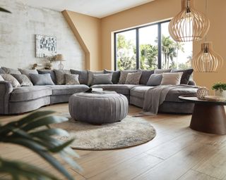 An L-shaped grey sofa by Sofology with beige wall paint decor, statement pendant ceiling lights, dark wooden table, round rug and cylindrical footstool