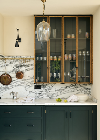 glass fronted cabinets with bamboo-like wooden lining above a dark green kitchen cabinet and a veined marble backsplash and counter