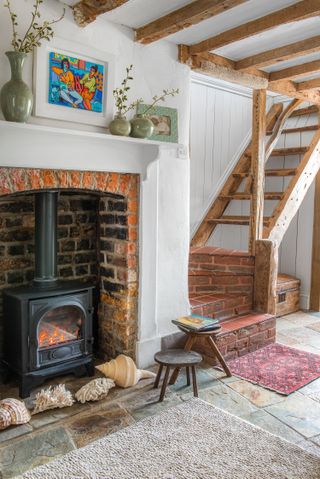 snug with fireplace with large shells and woodburner with small wooden stools bright painting above mantelpiece and stairs in background