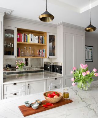 White fitted kitchen with mirror behind work top and open wooden shelves, and kitchen island with marble top, decorated with books, flowers, and wooden tray on island with cooking ingredients