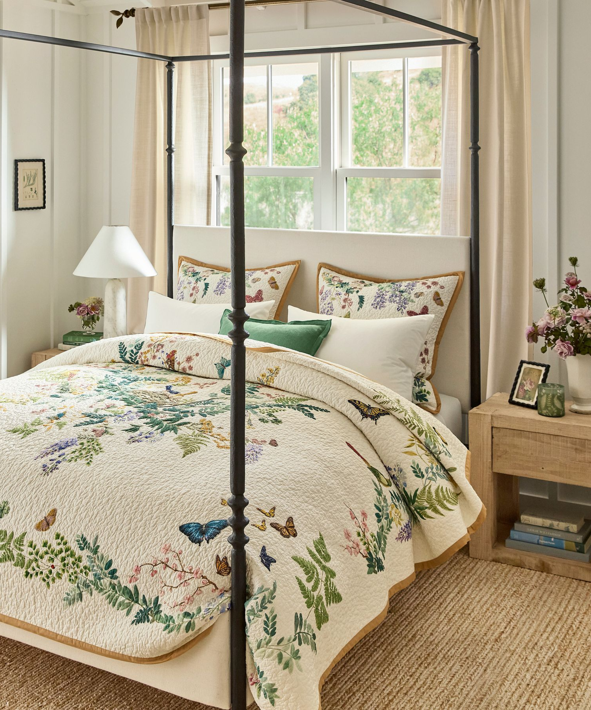 a cozy bedroom with a four-poster bed, a spring-themed quilt with flowers, ivy, butterflies, and birds on the bed, a small night stand with pictures and flowers, and a set of windows behind the bed with drapes in front of it