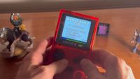 Hands hold a transparent red Game Boy Color displaying "Minicraft 3D." Nearby are figurines on a wooden surface, creating a nostalgic gaming vibe.