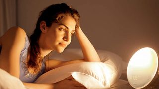 A woman lies in bed smiling at a Philips sunrise alarm clock which is glowing on her bedside table