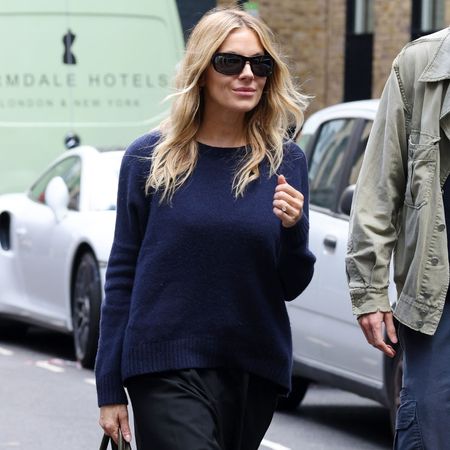 Sienna Miller steps out in London wearing flat black knee-high boots from Dune. She styled them with a grey knit and black knee-length shorts.