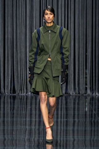 A female model wearing a green and blue striped zip up jacket, a green knee high dress and green high heel shoes walking down a runway.