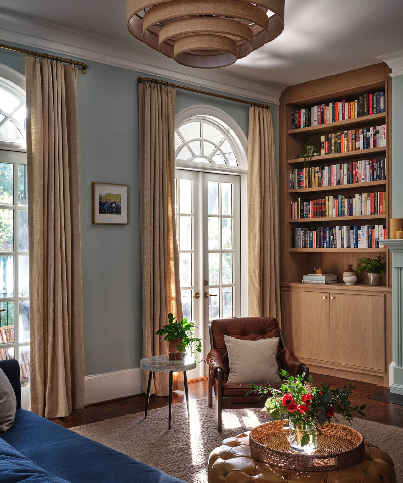 A light blue living room with wooden built-in bookshelves, a brown armchair, and large floor-to-ceiling white arched windows