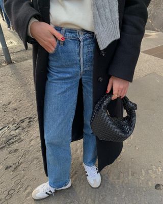 Woman wearing long, mid-wash blue jeans with white sneakers, black purse, white shirt, and long black coat from the waist down.
