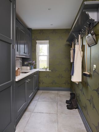A utility room with dark green cabinets and green motif wallpaper with coat hooks and a small window