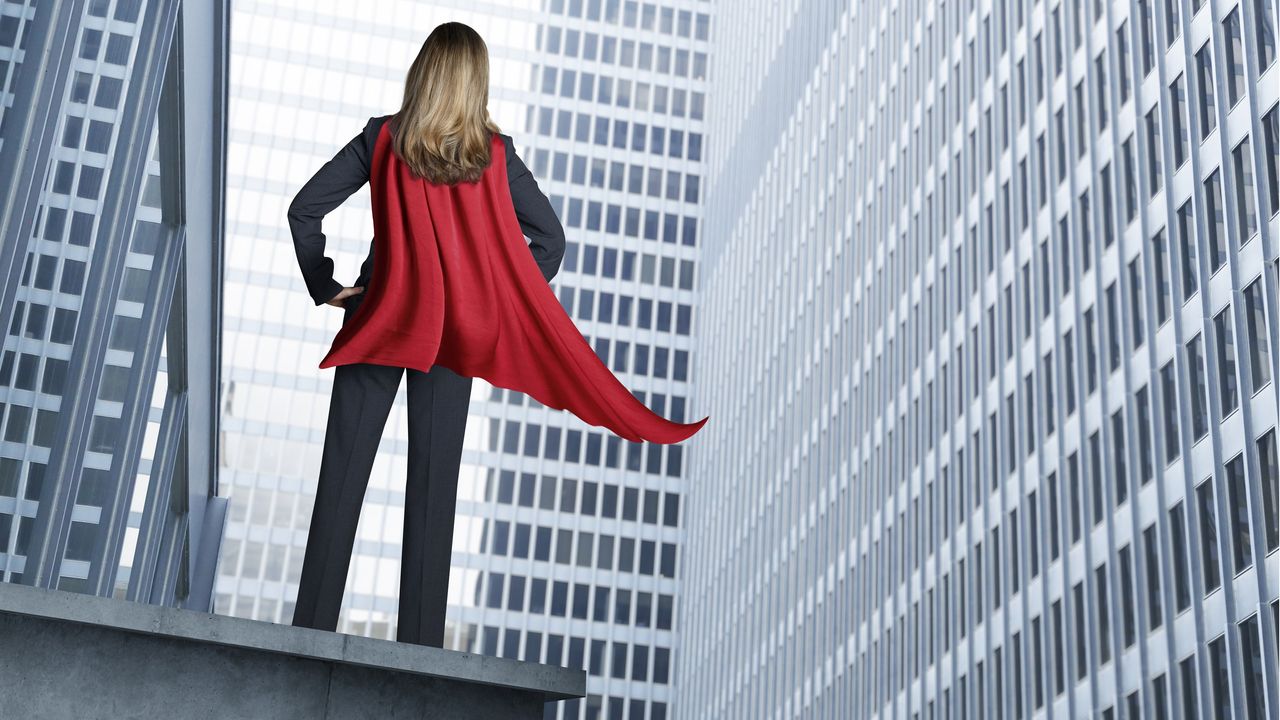 A woman wearing a red cape and business suit stands on the roof of a high-rise looking out at other high-rises.