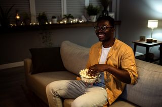 A man watching a movie in the evening while sitting on the sofa eating popcorn.