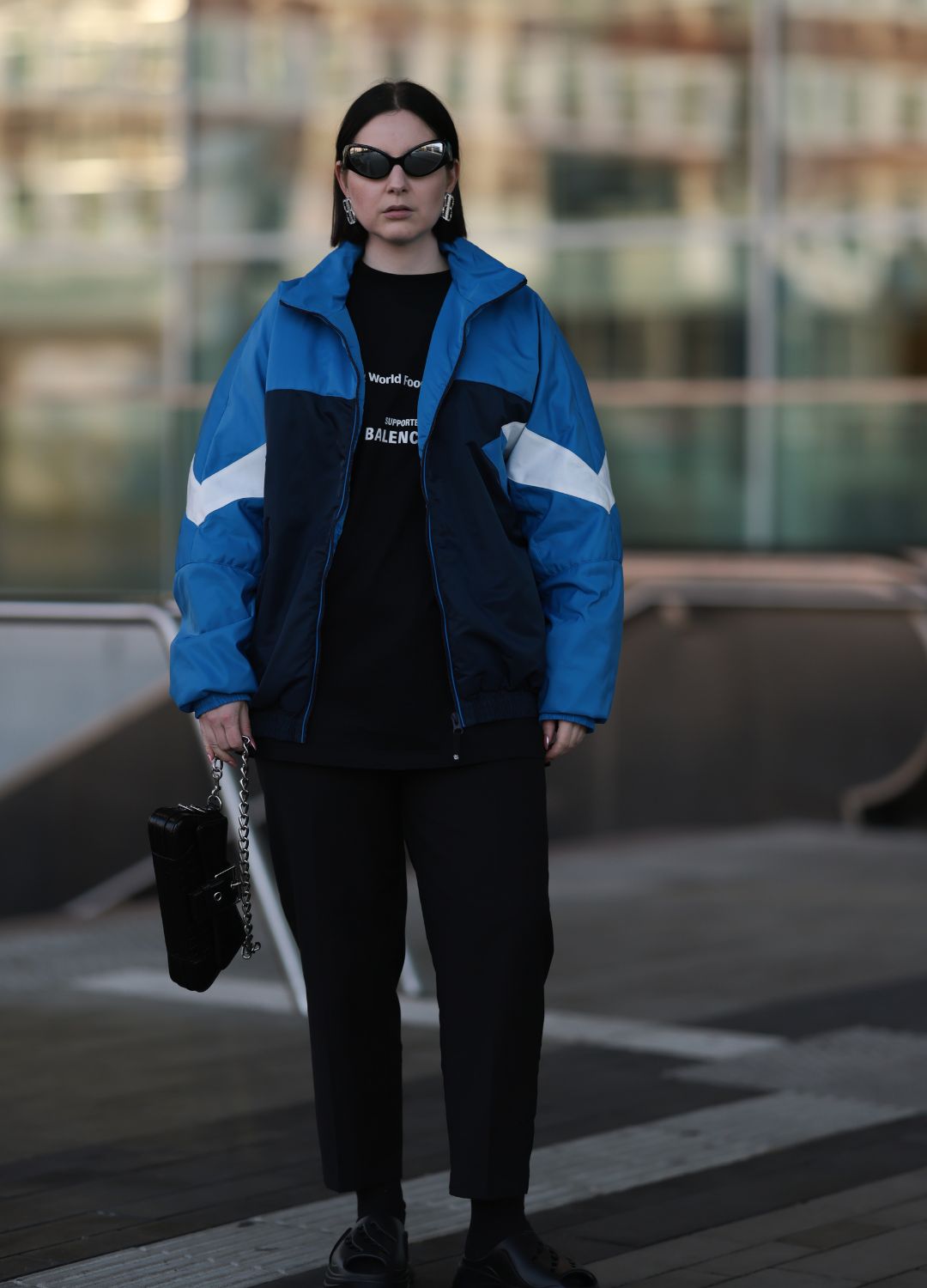 Maria Barteczko seen wearing Bershka black oval swift futuristic sunglasses, Balenciaga silver brass rhinestones logo earrings, Zara blue / white / black retro nylon tracksuit jacket, Balenciaga black logo cotton shirt, COS black classic pants, Adidas by StellaMcCartney black slipper clogs, Balenciaga black leather Lindsay chain bag, on March 18, 2023 in Dusseldorf, Germany