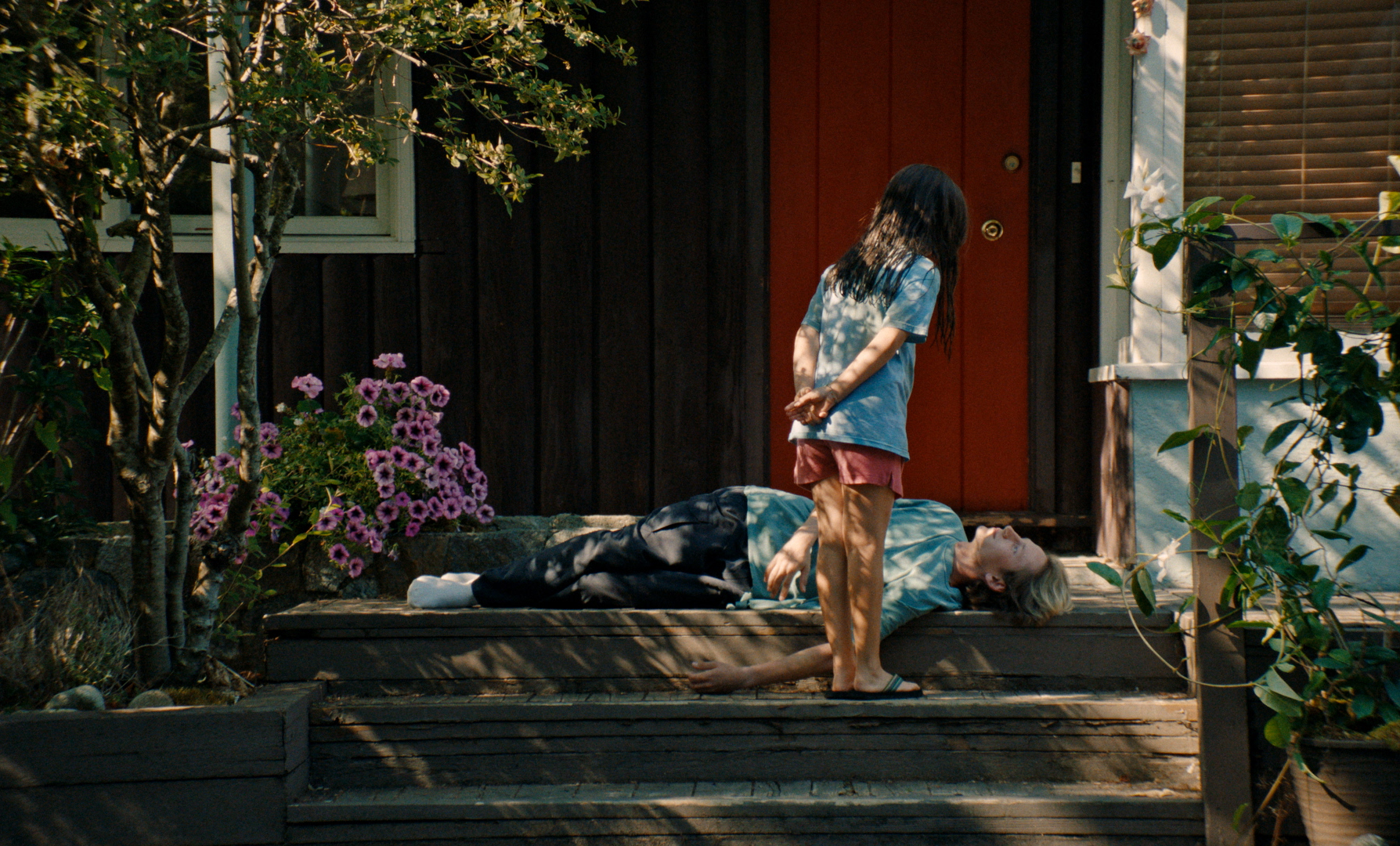 a little girl looks over a teenage boy lying on a front porch in the movie blue heron