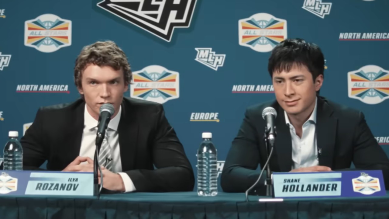 From left to right Ilya talking into a mic and Shane looking forward during a press confrence in Heated Rivalry. 